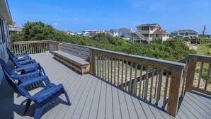 Een houten terras met blauwe stoelen en een bankje, met uitzicht op een strand met huizen op de achtergrond.