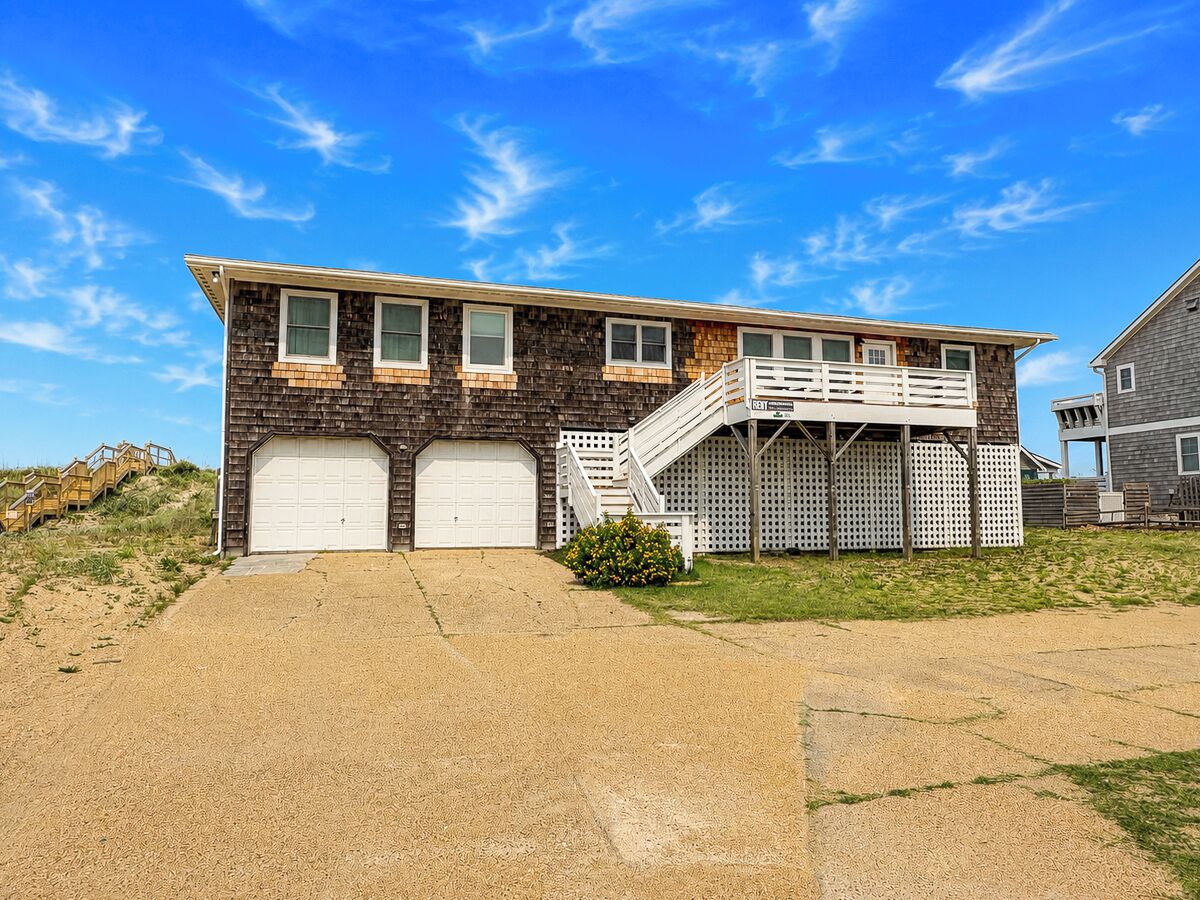 A two-story house with a garage, a staircase leading to a deck, and a sandy driveway.