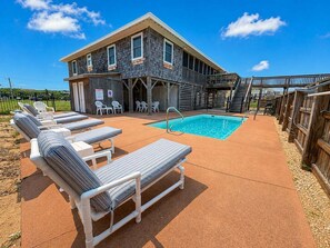 A pool area with lounge chairs, a pool, and a building with a covered deck. - Captain's R&R - 5 BR Oceanfront Home w/Private Pool & Hot Tub - Dogs Allowed (Southern Shores)