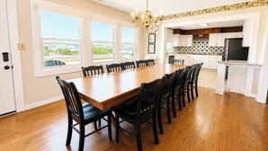 A spacious dining room with a large wooden table and chairs, a chandelier, and a view of the outside through large windows. - Captain's R&R - 5 BR Oceanfront Home w/Private Pool & Hot Tub - Dogs Allowed (Southern Shores)