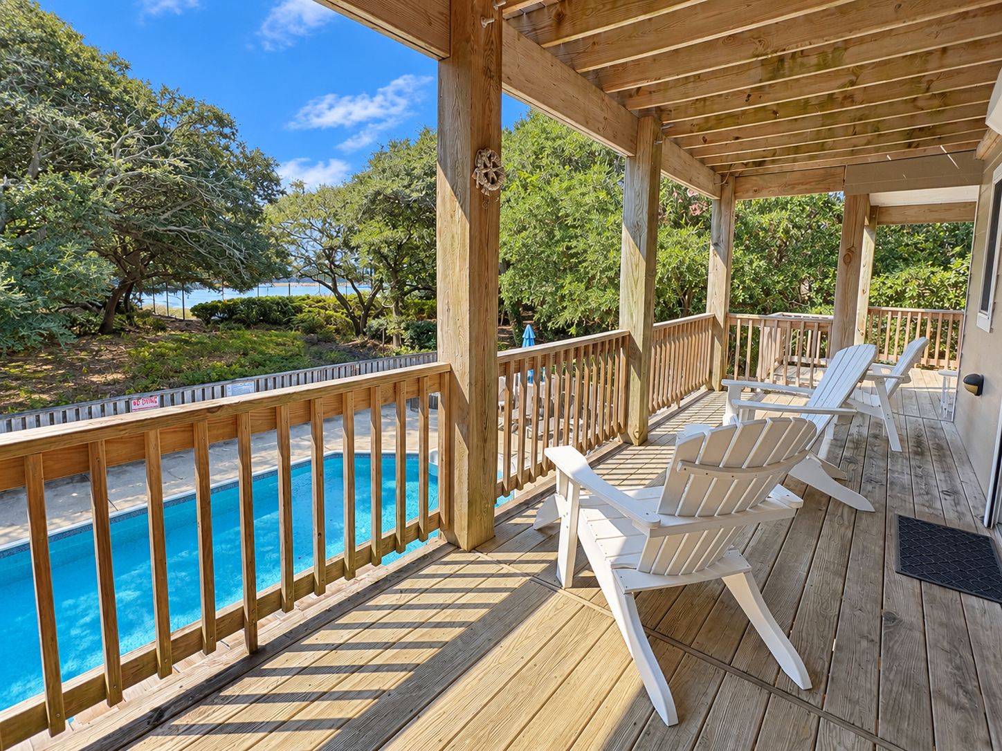 A wooden deck with a pool, white chairs, and a view of trees and water.