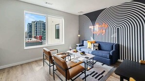 Una sala de estar moderna con un sofá, una mesa de centro, sillas y vistas a edificios a través de un ventanal grande.