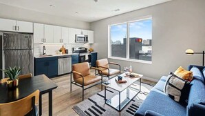 Una cocina moderna con armarios blancos, un microondas y un refrigerador. Hay un comedor con mesa y sillas, y un sofá con vista a la ciudad.