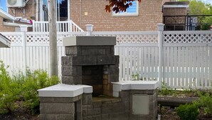 A stone-built outdoor fireplace with a white mantel, surrounded by a white picket fence, set in a garden with greenery and a brick house in the background. - Room-vermaurel Sweet Lodge (Bathurst)