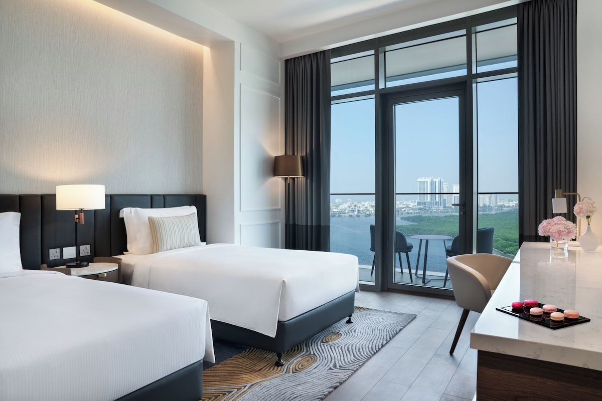 A hotel room with two beds, a desk, and a view of a cityscape through large windows.
