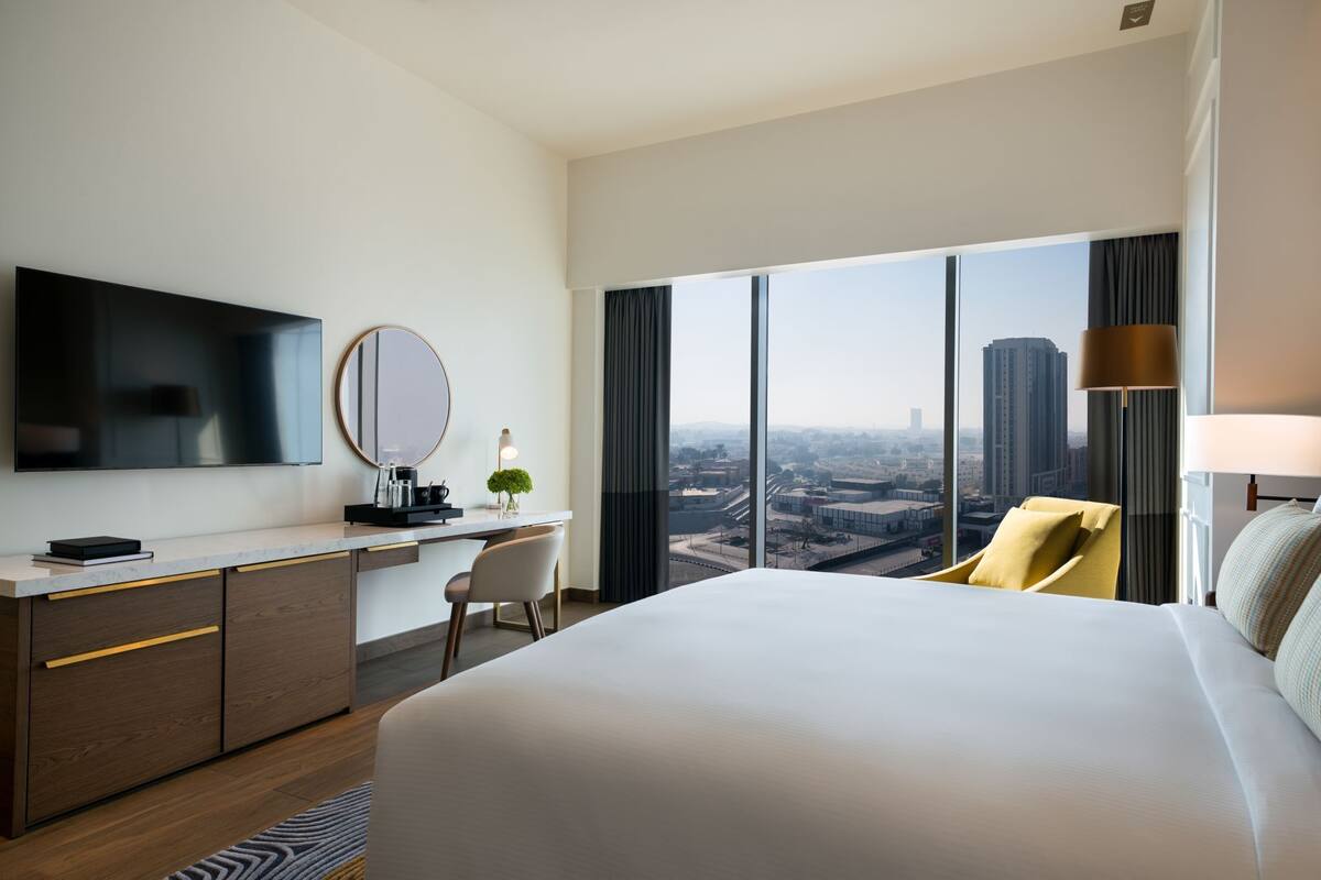 A hotel room with a large bed, a desk with a chair, a television, a round mirror, and a view of a cityscape through large windows.