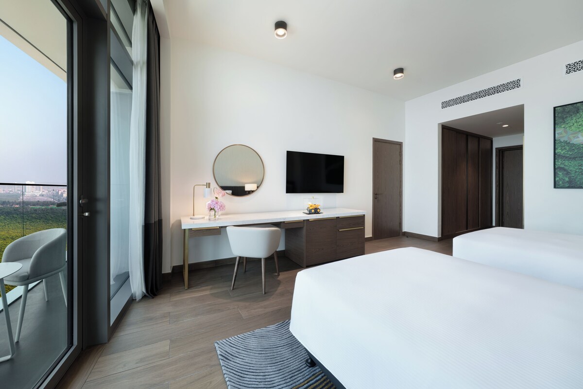 A modern hotel room with a large bed, a desk with a chair, a flat-screen TV, and a large window offering a city view.