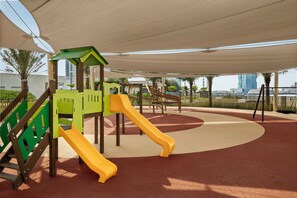 A playground with a slide and climbing structures, surrounded by a covered area with a view of a cityscape. - Rotana Ras Al Khaimah - The Mangroves (Ras Al Khaimah)