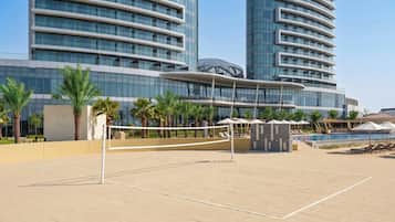 Una cancha deportiva con red de voleibol, palmeras y un edificio moderno con ventanas de vidrio reflectante al fondo.
