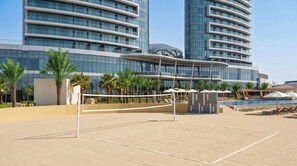 A sports court with a volleyball net, palm trees, and a modern building with reflective glass windows in the background. - Rotana Ras Al Khaimah - The Mangroves (Ras Al Khaimah)