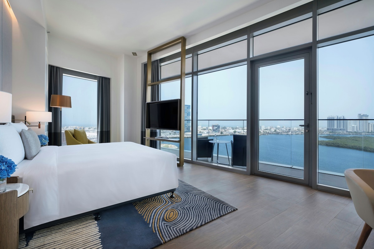 A modern hotel room with a large bed, a flat-screen TV, a dining area with a view of the water, and a balcony with a cityscape view.