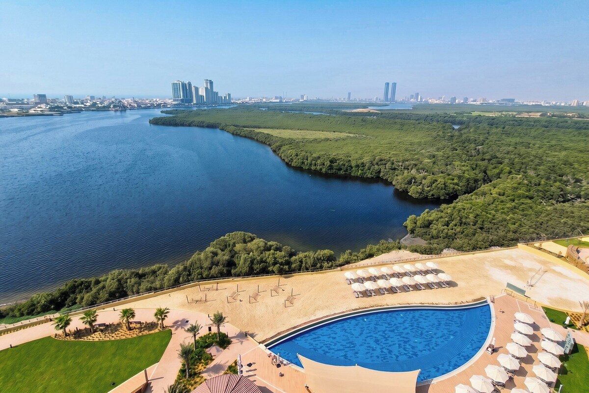 A resort with a swimming pool, beach area, and views of a river and city skyline.
