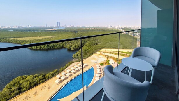 A balcony with a view of a river, a swimming pool, and a cityscape in the distance. - Rotana Ras Al Khaimah - The Mangroves (Ras Al Khaimah)