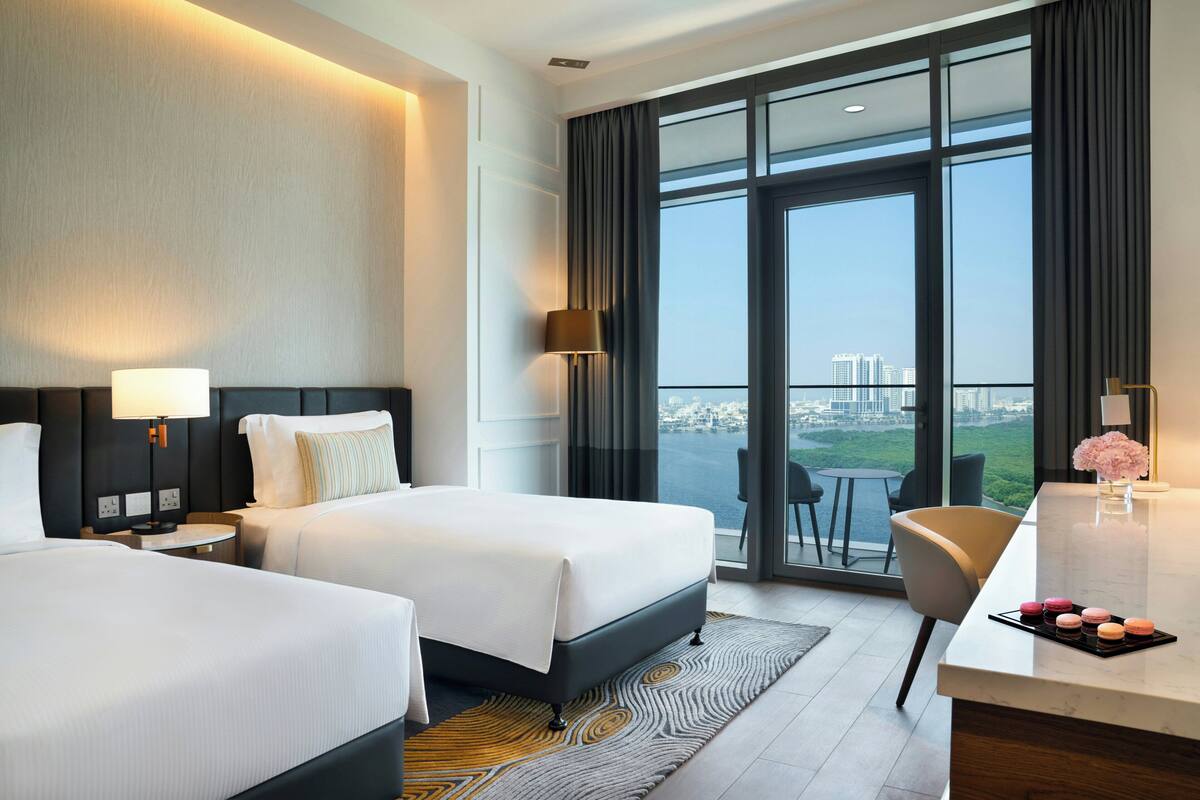 A hotel room with two beds, a desk, a chair, a view of the city, and a balcony.