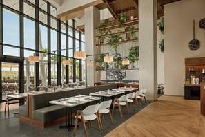 A modern restaurant interior with high ceilings, large windows, and a variety of seating options including tables and a long bench. - Rotana Ras Al Khaimah - The Mangroves (Ras Al Khaimah)