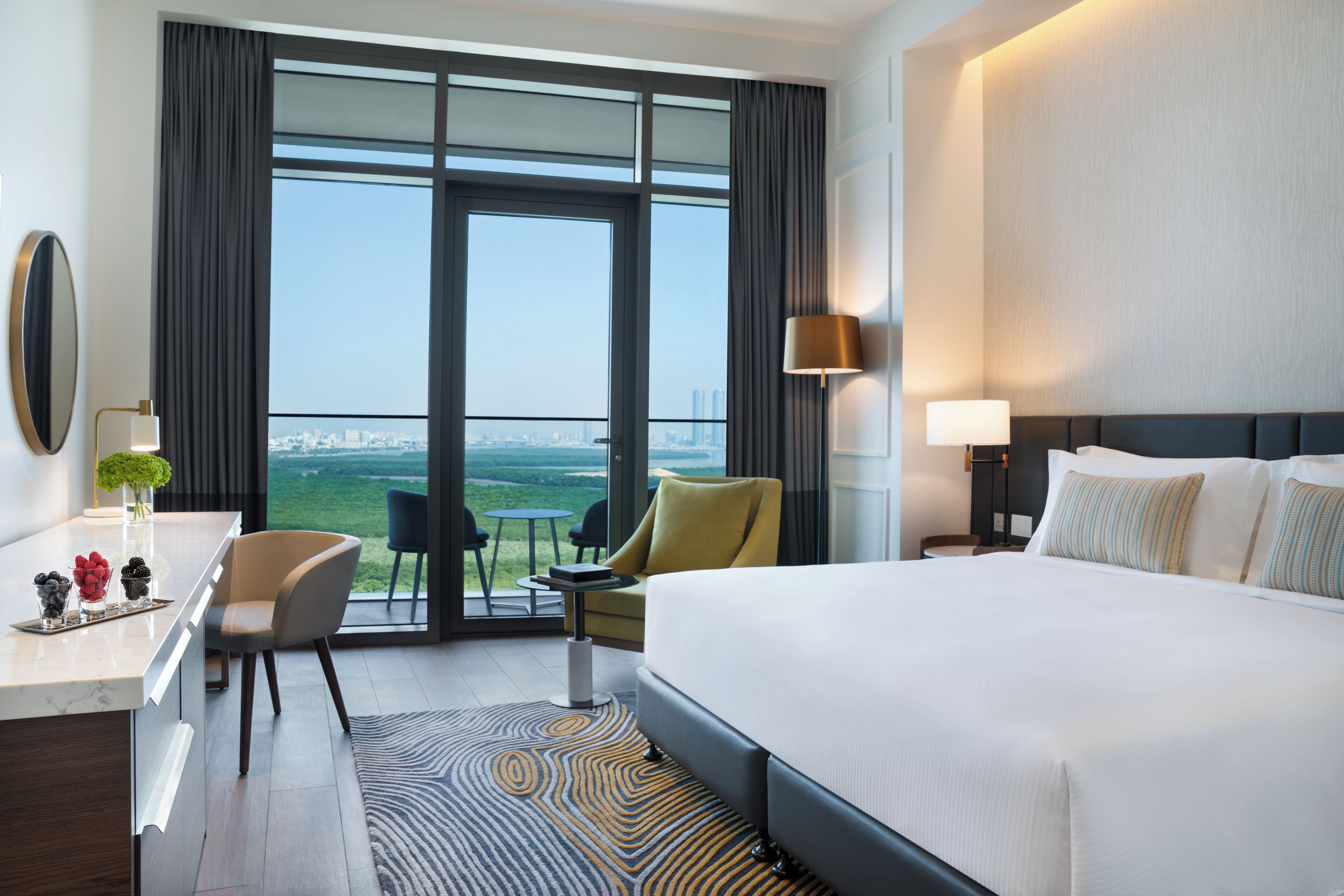 Habitación de hotel con una cama grande, un escritorio con espejo, una silla, una mesita y vistas a la ciudad a través de la ventana.