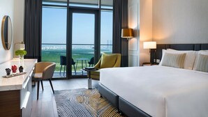 A hotel room with a large bed, a desk, a chair, a small table, and a view of the city through large windows. - Rotana Ras Al Khaimah - The Mangroves (Ras Al Khaimah)
