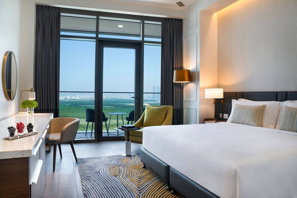 A hotel room with a large bed, a desk, a chair, a small table, a lamp, and a view of the city.