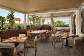 An outdoor dining area with tables and chairs, surrounded by greenery and a view of palm trees. - Rotana Ras Al Khaimah - The Mangroves (Ras Al Khaimah)