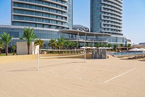 A beach volleyball court with a net, palm trees, and a pool area with lounge chairs and umbrellas. - Rotana Ras Al Khaimah - The Mangroves (Ras Al Khaimah)