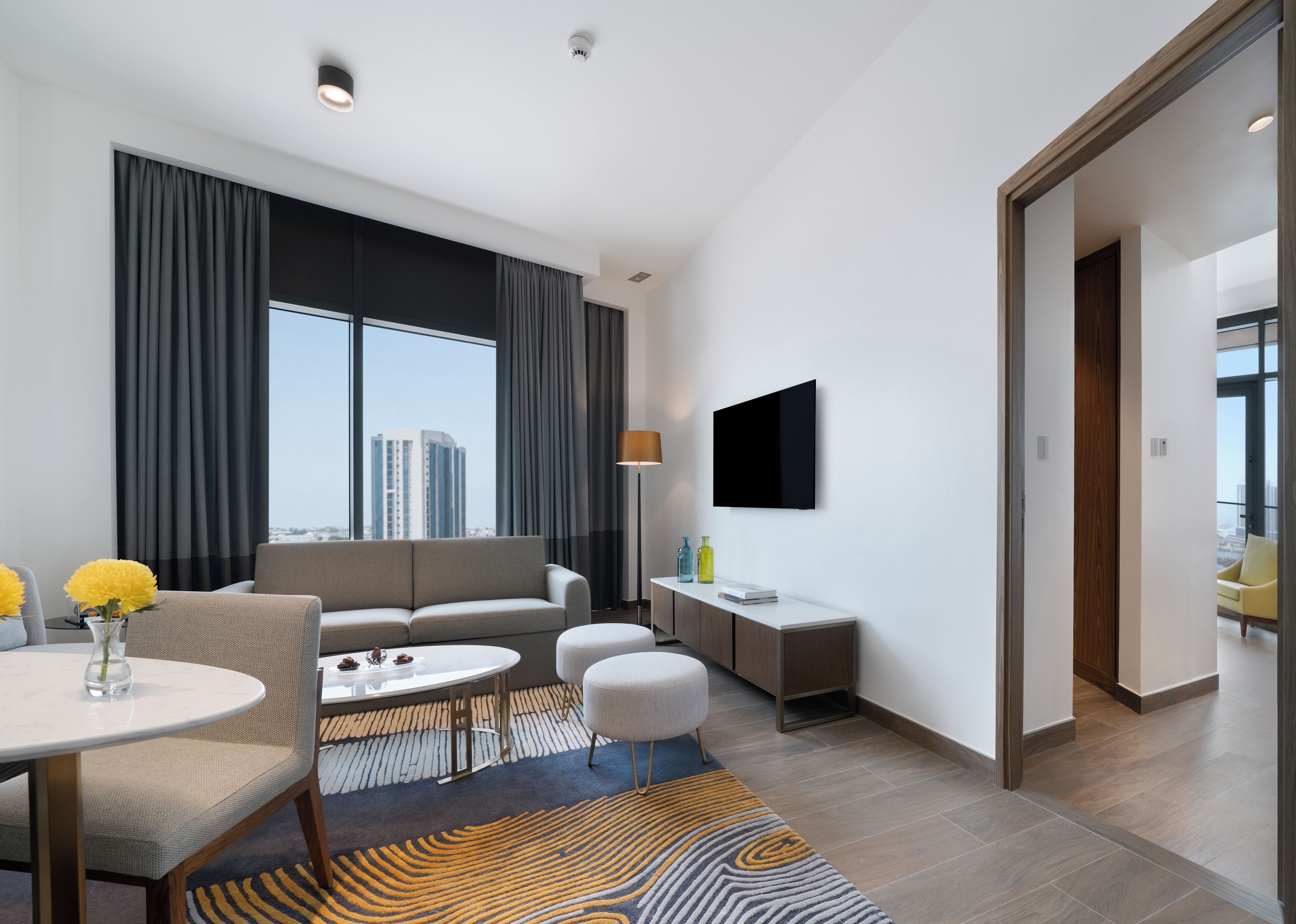 Una habitación de hotel moderna con sofá, una mesa de comedor, televisor montado en la pared y vistas a la ciudad.