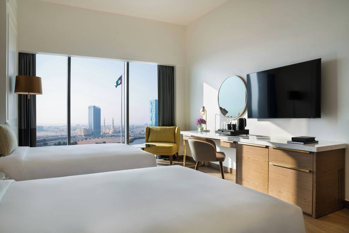 A hotel room with two beds, a desk, a chair, a TV, and a large window with a city view.