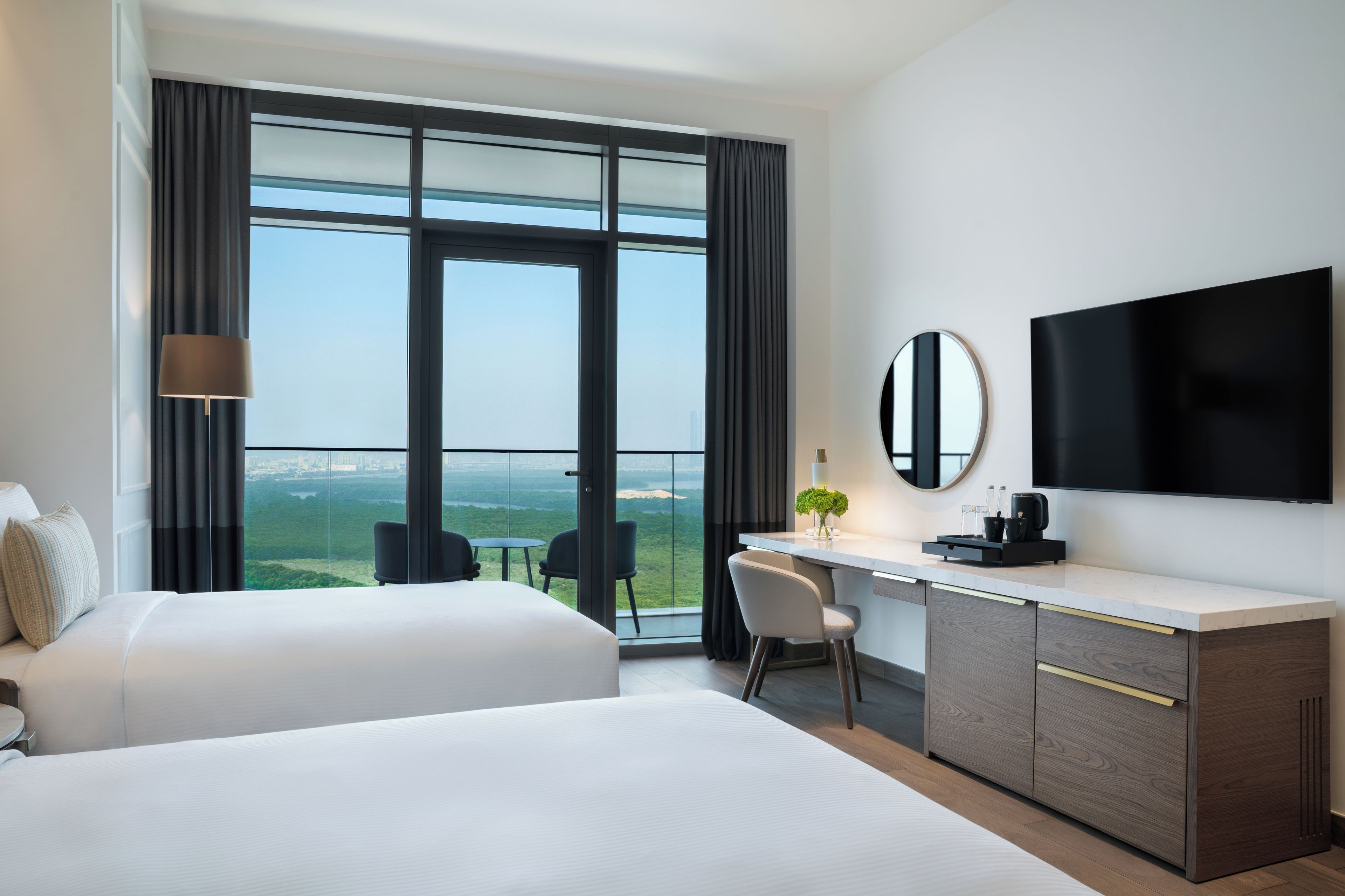 A hotel room with two beds, a desk, a chair, a TV, and a large window with a view.
