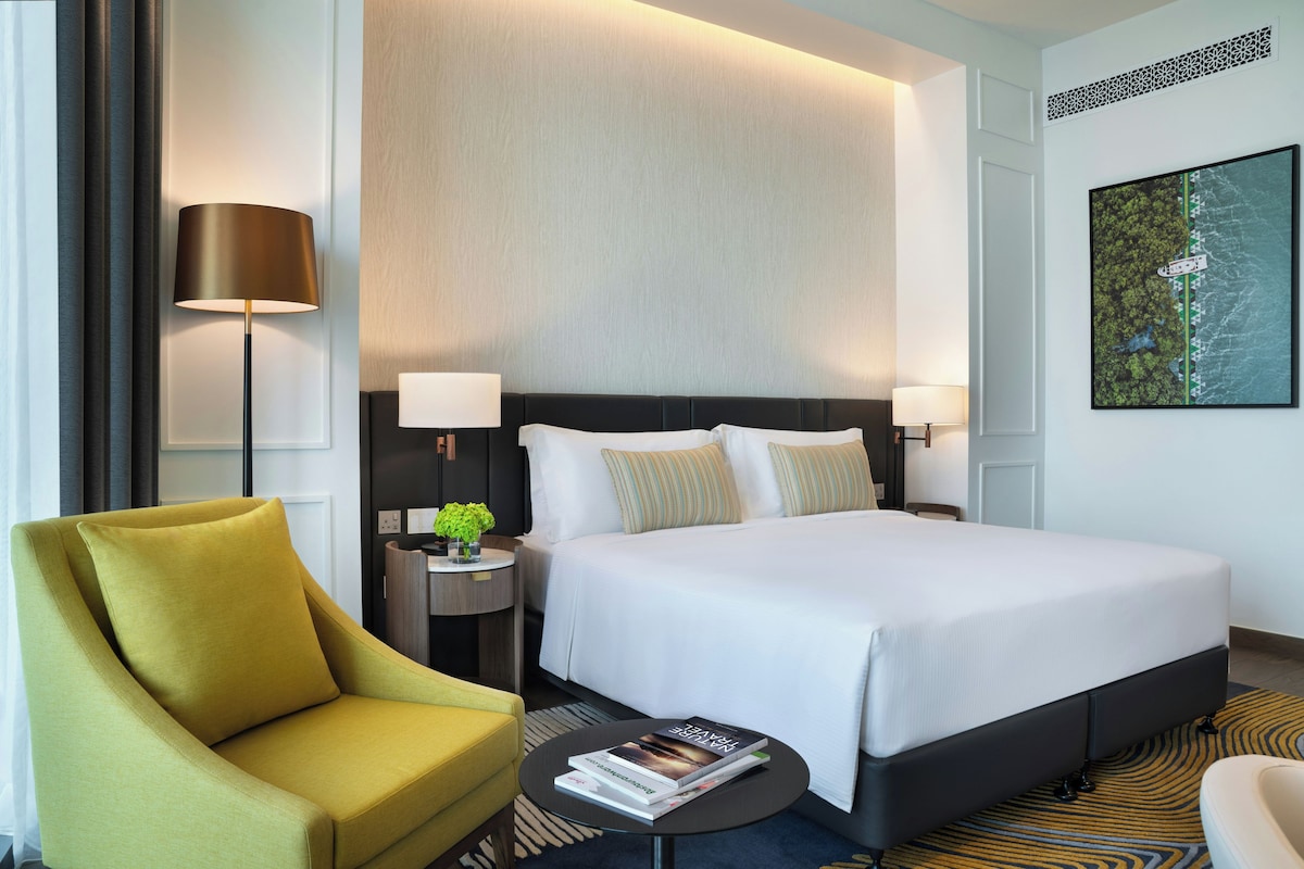 A hotel room with a bed, a chair, a nightstand, a lamp, a small table with a book, and a wall-mounted artwork.