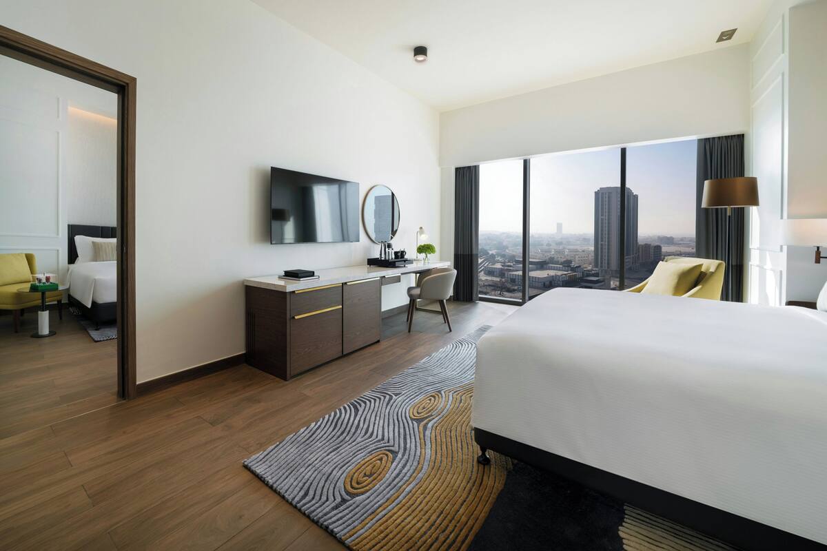 A modern hotel room with a large bed, a desk, a chair, a television, and a view of the cityscape.