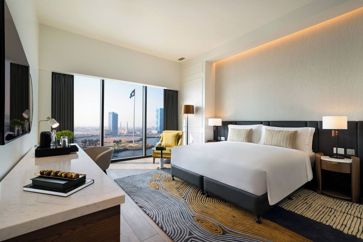 A modern hotel room with a large bed, a desk, a chair, and a view of the cityscape through large windows.