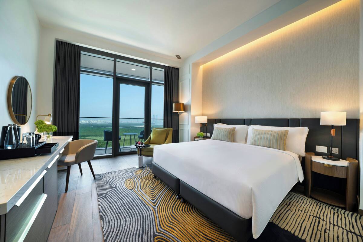 A modern hotel room with a large bed, a desk, a chair, a nightstand, and a view of the outdoors.