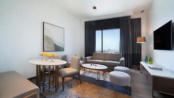A modern hotel room with a sofa, two armchairs, a dining table, a TV, and a large window with a city view.