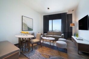 A modern hotel room with a sofa, two armchairs, a dining table, a TV, and a large window with a city view. - Rotana Ras Al Khaimah - The Mangroves (Ras Al Khaimah)