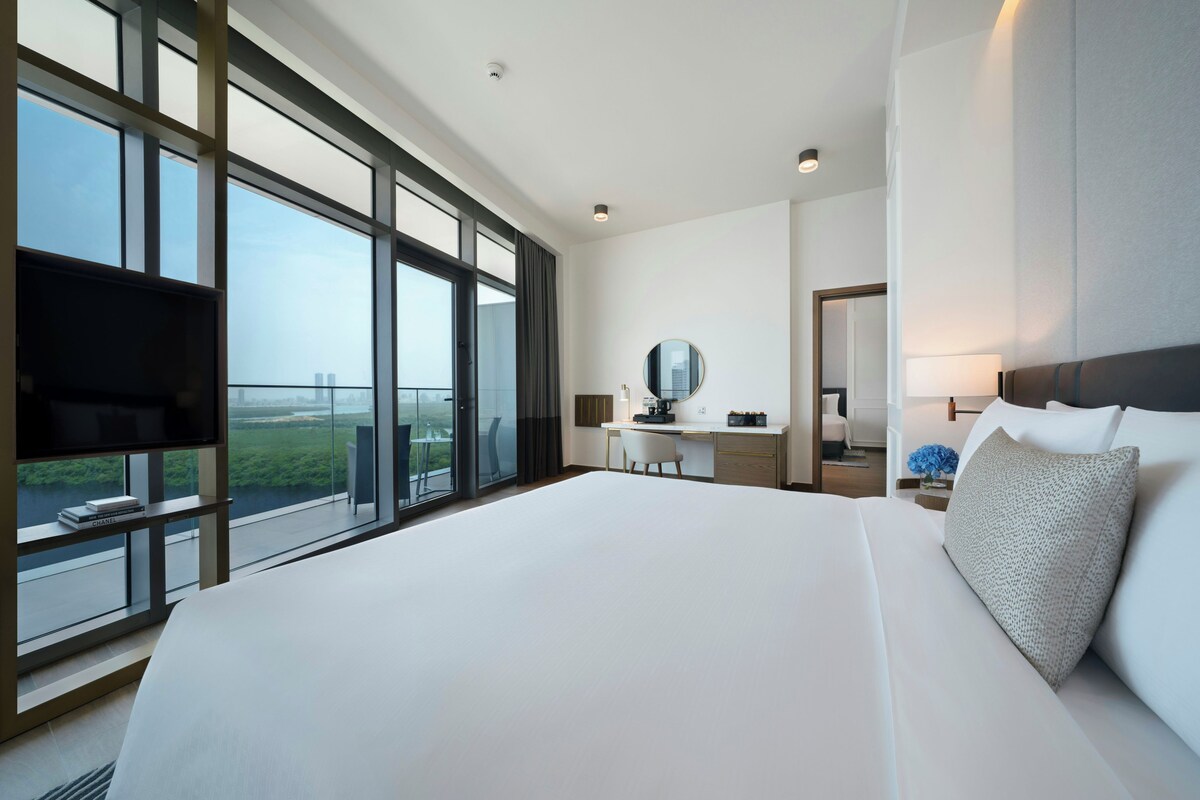 A modern hotel room with a large bed, a desk, a chair, a television, and a view of the city.