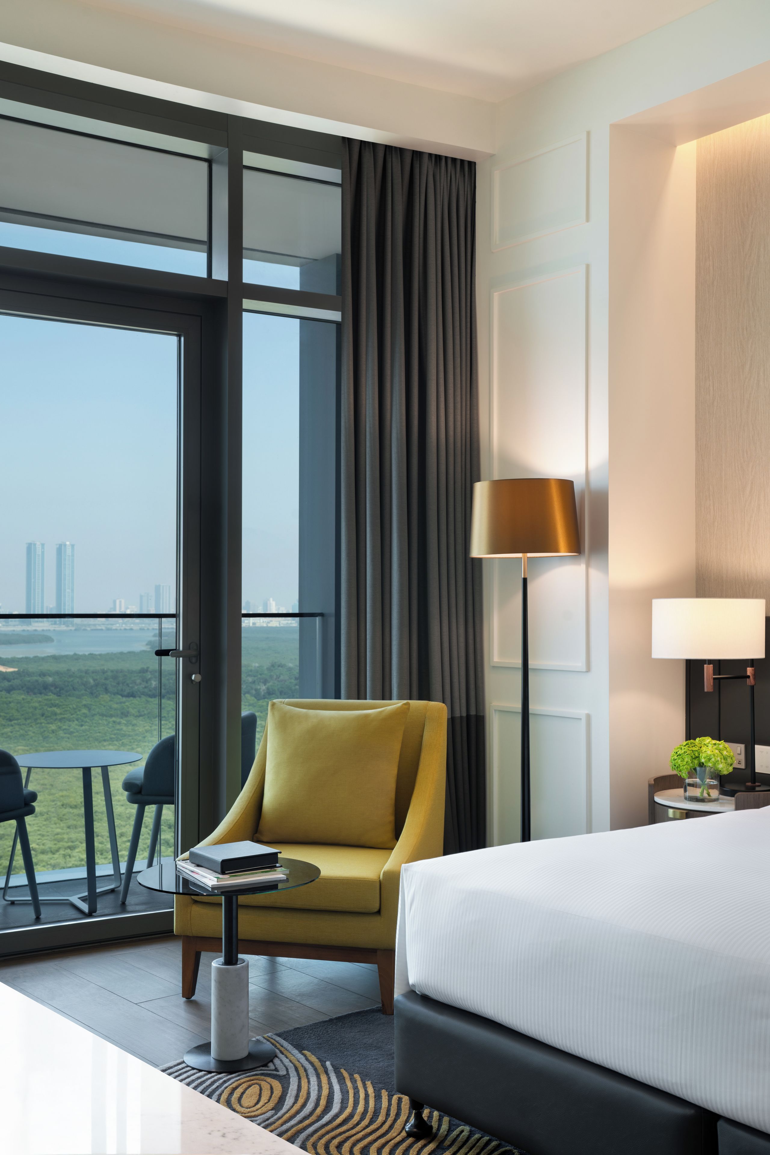 Ein Hotelzimmer mit Bett, einem gelben Sessel, einem kleinen Tisch, einer Lampe und Blick auf die Stadt durch große Fenster.