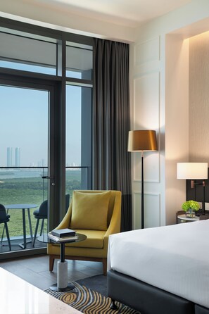 A hotel room with a bed, a yellow armchair, a small table, a lamp, and a view of the cityscape through large windows.