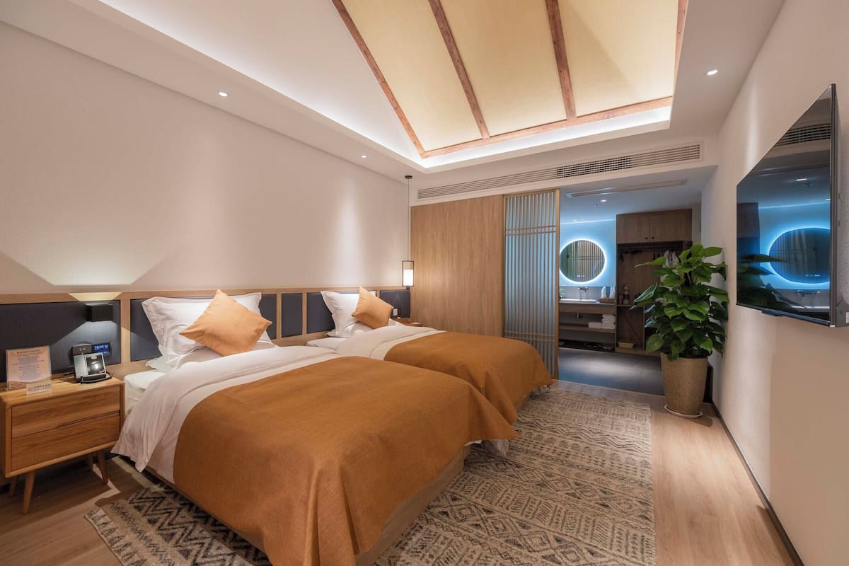 Una habitación de hotel moderna con dos camas, una mesita de noche, un escritorio, un espejo grande y una planta en maceta.