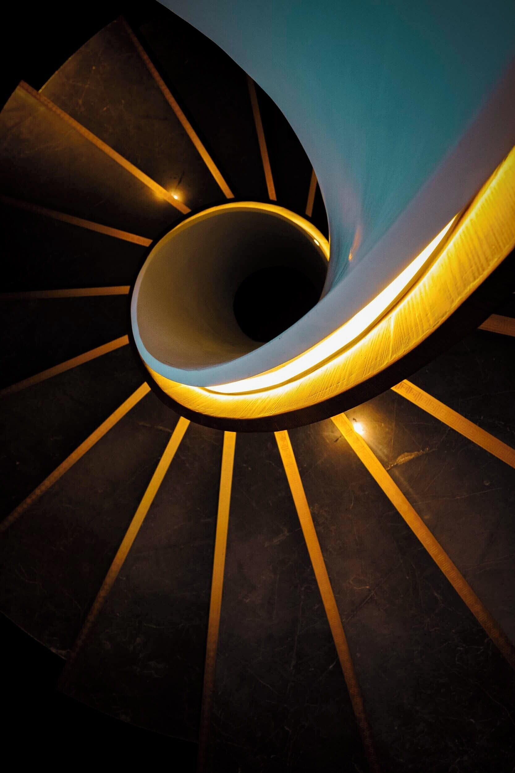 A spiral staircase with a central illuminated handrail and a dark, reflective floor.