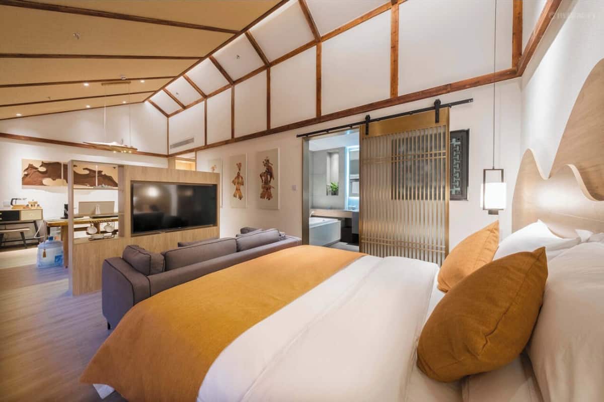 Una habitación de hotel moderna con una cama grande, un sofá, un televisor y una puerta corredera hacia un baño.