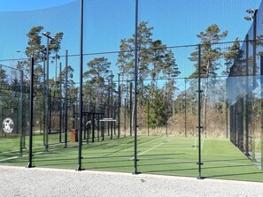 Una cancha de tenis con una red, rodeada por una valla y árboles al fondo.