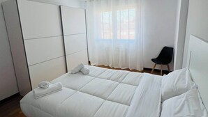Una cama bien hecha con sábanas y almohadas blancas, una silla negra y una ventana con cortinas traslúcidas.