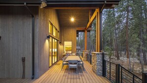 A wooden deck with a dining area, stone walls, and a view of a forest. - Luxe Caldera Escape w/ Forest Views, Private Hot Tub, Sauna, Game Room, Seasonal Pool, Bikes & More! (Bend)