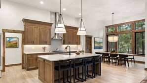 A modern kitchen with wooden cabinets, a large island with bar stools, pendant lighting, and a view of trees through large windows. - Luxe Caldera Escape w/ Forest Views, Private Hot Tub, Sauna, Game Room, Seasonal Pool, Bikes & More! (Bend)