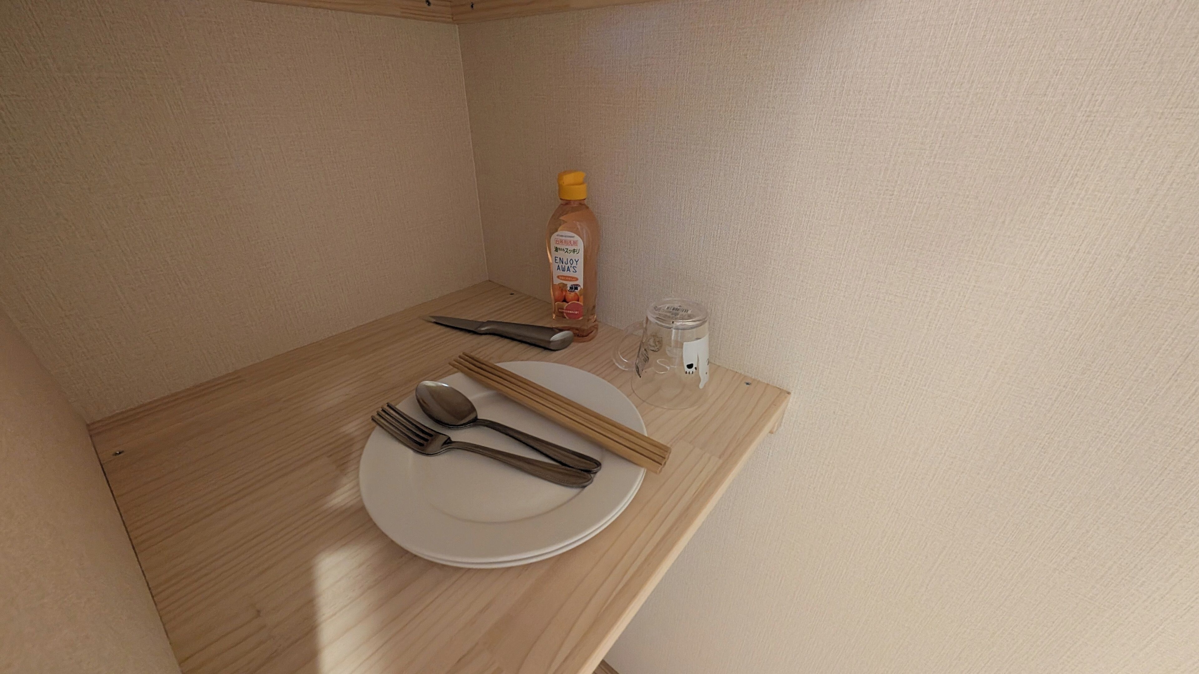 A small wooden table with a white plate, chopsticks, a spoon, a fork, a bottle of sauce, and a small glass jar.