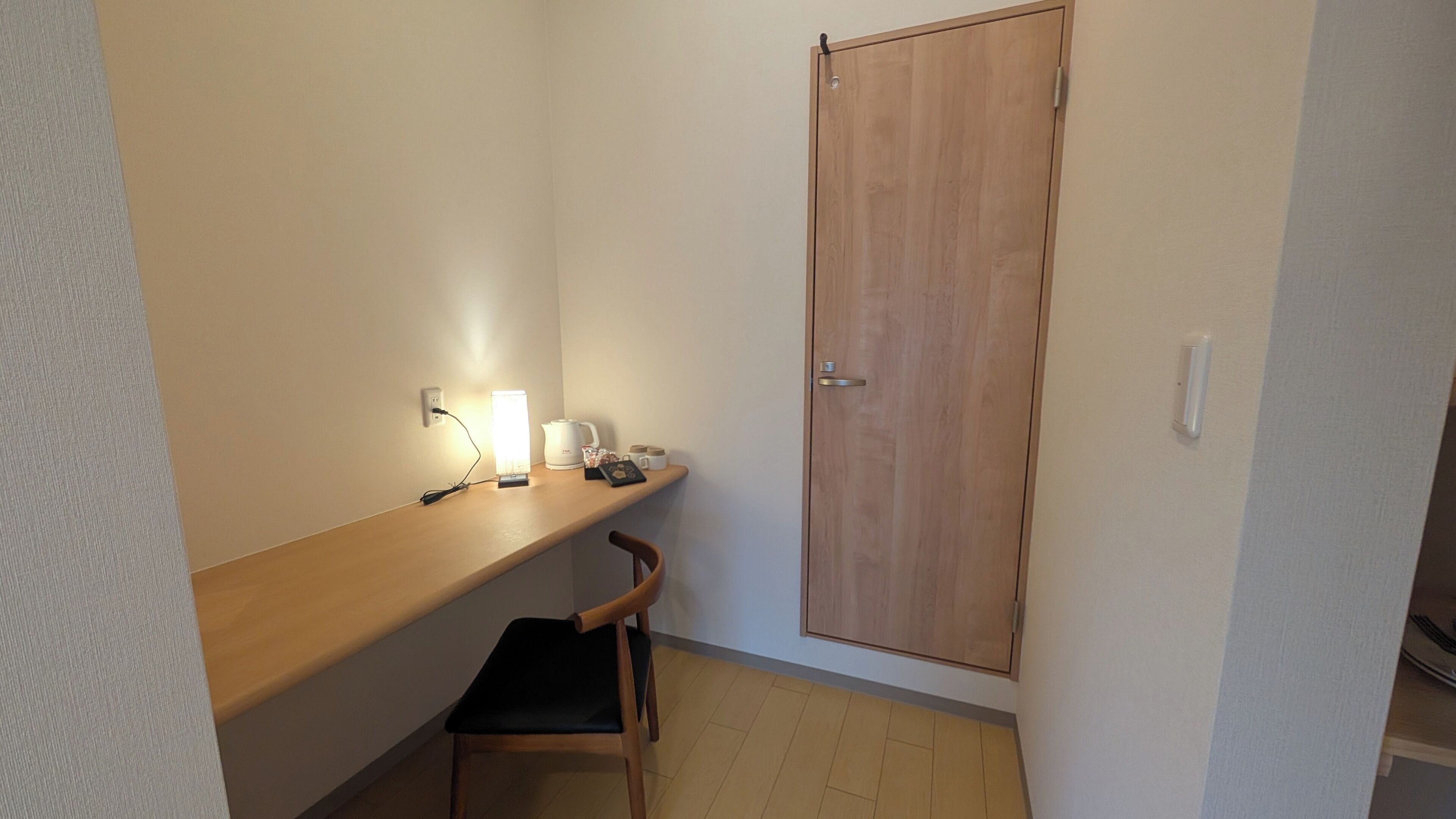 A small, narrow room with a wooden desk, a chair, a wooden door, and a light fixture on the wall.