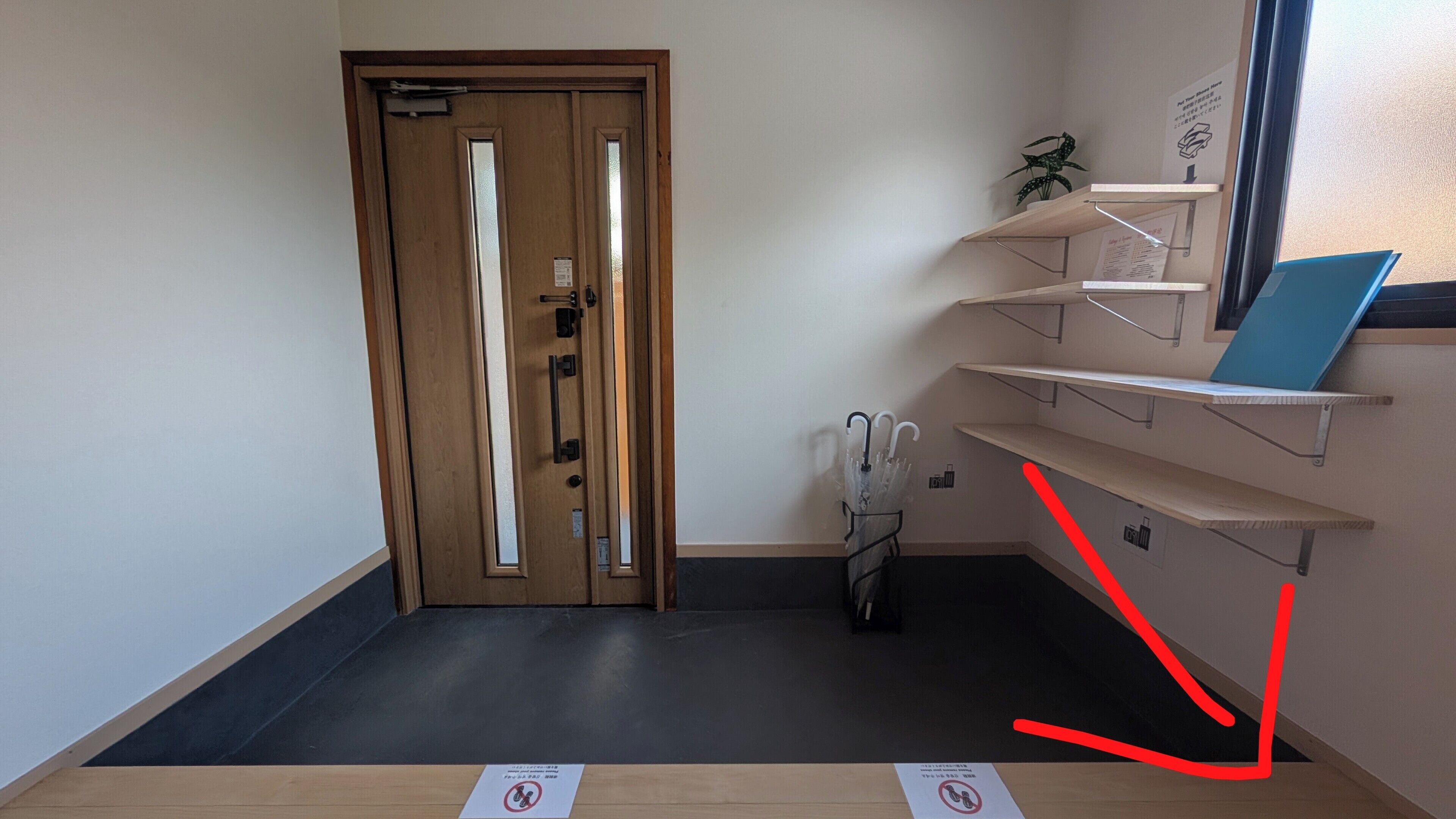 A hotel room with a wooden door, a black floor, and a window with a plant on the sill. There are two signs on the floor, one with a no-smoking symbol and another with a no-entry symbol.