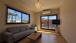 A modern living room with a grey sofa, a wooden coffee table, a potted plant, a TV on a stand, and large windows offering a view of buildings and a fence. - Dream Stays SORA (Izumisano)