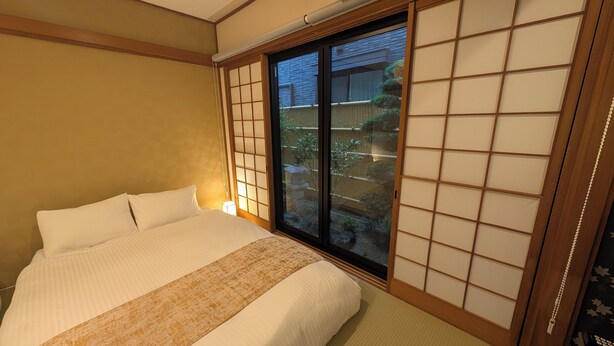 A traditional Japanese-style bedroom with tatami flooring, a low bed with white bedding, and a sliding glass door leading to a garden. - Dream Stays SORA (Izumisano)