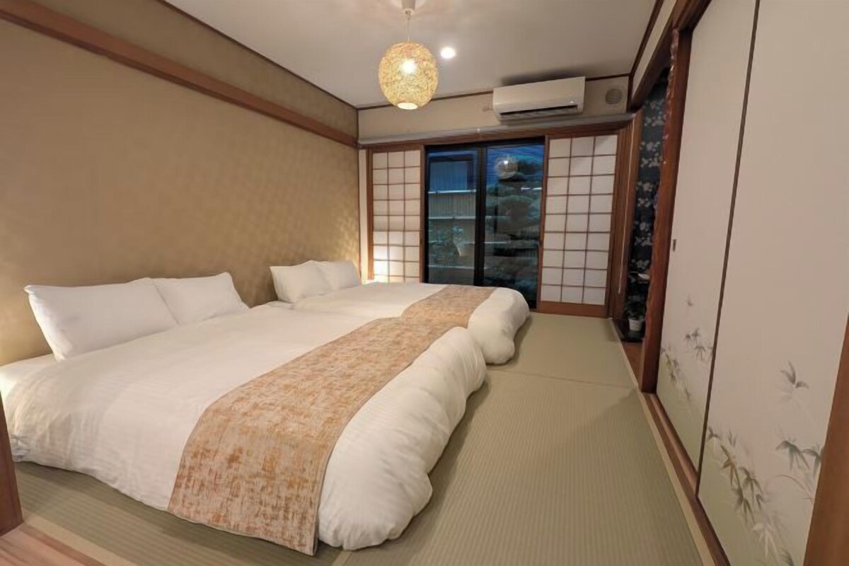 A traditional Japanese-style bedroom with tatami flooring, a large bed with white bedding, a hanging light fixture, and sliding shoji doors.