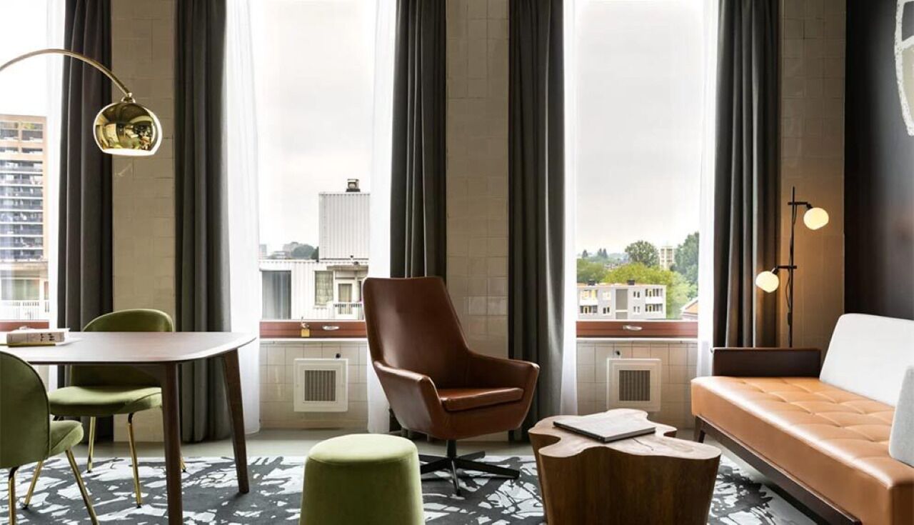 Quarto de hotel moderno com uma grande janela, uma poltrona de couro marrom, uma mesa de centro de madeira, uma poltrona verde e um sofá.